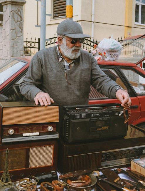 An elderly man with a white beard, wearing a gray flat cap, sunglasses, a light gray sweater, and a checked shirt, is standing outdoors behind a red vehicle. He is holding a string of brown beads in his right hand while his left hand rests on a vintage portable radio placed on a partially opened vehicle door. In front of him, on the vehicle's surface, there are various objects, including a small Eiffel Tower replica, several pieces of jewelry, and other miscellaneous items. Behind and around him, there are bags and boxes, suggesting a street sale or private collection setup. The background features a stone wall and a building with cream-colored walls and a metal fence. The scene appears to be in an outdoor urban environment, possibly during daytime, with natural lighting illuminating the scene clearly, highlighting the textures of the clothing, objects, and surrounding environment. This setup aligns with the concept of on-site clearance or independent collection of second-hand items, similar to what Rubbish Removal Bow might handle in waste removal services for traders or private individuals. The overall scene emphasizes a casual yet organized display of goods, indicating a transaction or exchange related to rubbish removal or disposal of unwanted items.