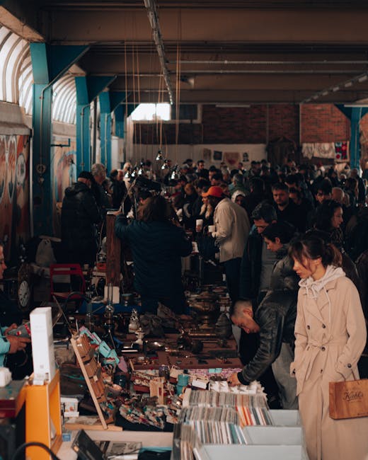 Inside a large market hall, numerous traders and visitors browse and purchase various items displayed on tables. The scene is bustling with activity, featuring diverse individuals dressed in casual apparel, with some wearing jackets and coats suitable for indoor environments. The tables are filled with an array of goods, including textiles, books, and miscellaneous household items, which are arranged in a somewhat cluttered fashion typical of market stalls or second-hand sales. The interior of the hall has a high, corrugated metal roof with exposed beams and hanging light fixtures, and the walls are a mix of brick and painted surfaces. Natural light filters in through windows near the ceiling, illuminating the vibrant atmosphere. Visible on the side are some discarded or leftover packaging materials, while the overall scene suggests an organized yet busy environment where some traders might be involved in the collection or disposal of waste materials associated with market trade, aligning with the services offered by Rubbish Removal Bow for external or on-site waste management related to trading activities.