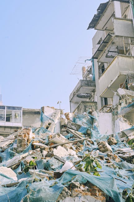 The image shows a pile of mixed rubble and debris in an outdoor urban setting, likely part of a demolition or clearance site. In the foreground, there are scattered bricks, concrete fragments, wooden planks, and pieces of metal, some of which are partially covered with blue tarpaulin or plastic sheets. The debris appears to be a combination of construction waste materials with varying textures and colours, including weathered reds, greys, and browns. Behind the rubble, there are partially visible sections of damaged or unfinished building structures, with exposed brickwork, plaster, and concrete surfaces. The surrounding environment includes a cityscape with high-rise residential buildings, featuring balconies, windows, and external cladding. Some areas show signs of damage or neglect, with dislodged materials and debris on the ground surface. The sky above is clear and bright, with natural daylight illuminating the scene. This type of scene is typical of sites where independent waste clearance or rubbish removal services, such as those provided by Rubbish Removal Bow, might be engaged to clear construction and demolition waste efficiently.