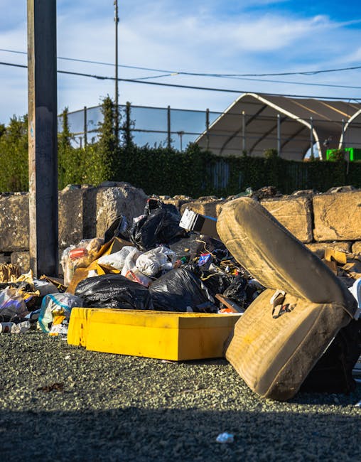 A close-up view of a pile of mixed household rubbish dumped on a gravel surface beside a stone wall, consisting of black plastic garbage bags, a yellow plastic container, an old, dirty beige car seat, and scattered debris. The black trash bags appear full and are partially torn, revealing paper and plastic waste inside. The yellow container is empty and lying flat on the ground, while the car seat is weathered with visible fabric wear and discoloration. In the background, there is a stone wall border, a metal pole, and a green hedge, with a sports court or similar recreational area enclosed by fencing and a canopy structure visible further back under a partly cloudy sky. The scene suggests an instance of unsanctioned rubbish disposal, highlighting the importance of professional waste management services like those offered by Rubbish Removal Bow, which specialise in private on-site clearance for domestic or commercial clients.