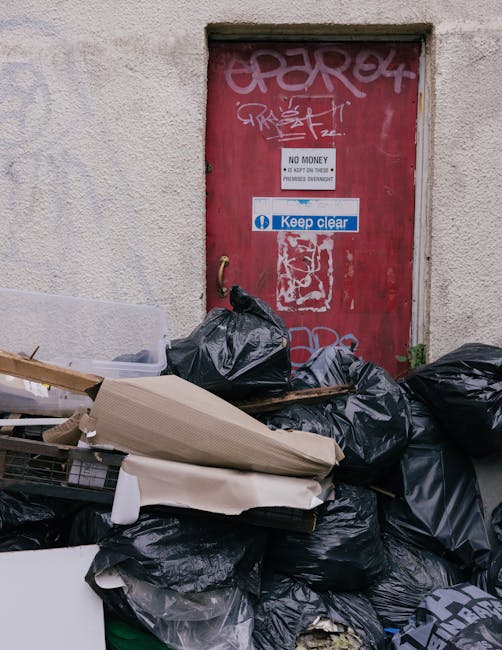 A collection of black garbage bags, some tied and others partially open, are piled in front of a red metal door set into a beige, textured wall. The bags appear to contain waste and miscellaneous discarded items. Among the debris, there are flattened cardboard boxes and a piece of white foam or packaging material. The door features multiple layers of graffiti in white and pink spray paint, along with two signs: one reading 'No Money Keep Out' and another blue and white sign stating 'Keep clear.' The scene is outdoors, possibly on a street or alleyway, with natural daylight illuminating the area. The surrounding wall has a rough, stucco finish, and the door frame is simple and unadorned. This scene illustrates an example of unmanaged rubbish accumulation which private waste removal services, such as those provided by Rubbish Removal Bow, can collect through on-site clearance or alternative waste handling methods, ensuring proper disposal especially in contexts where regular local authority rubbish collection may not be available or sufficient.