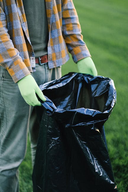 A person wearing a yellow and grey checkered shirt, grey trousers, and light green gloves is holding open a large black plastic rubbish bag, preparing to dispose of waste outdoors on a grassy area. The background shows a lush, green, well-maintained lawn, suggesting a park or garden setting. The person's hands are gripping the sides of the bag, which appears to be ready for collection or removal. The scene implies the activity of waste clearance, possibly as part of an individual or private rubbish removal effort, highlighting the importance of proper waste handling and disposal outside of local authority services. The lighting is natural, with daylight illuminating the scene evenly, creating a clear, detailed view of the materials and textures, such as the shiny, crinkled surface of the plastic bag and the fabric of the gloves. This image illustrates the process of on-site rubbish collection or pre-collection preparation, aligned with the principles of independent waste management and private disposal methods supported by companies like Rubbish Removal Bow.