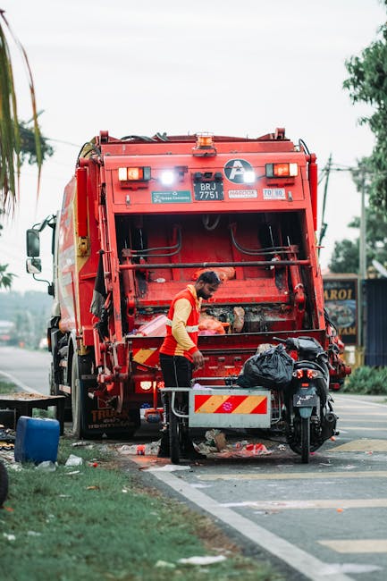 A worker wearing a red and yellow uniform is operating a large, red rubbish collection vehicle parked on the side of a street. The back of the vehicle is open, revealing mechanical components and a collection chamber designed for waste storage. The worker is managing rubbish or debris, which appears to include black plastic bags, some loose waste, and small fragments scattered on the ground nearby. The scene is outdoors with a road, some trees, and a few buildings visible in the background, indicating an urban or suburban environment. The street has a painted line marking the edge of the traffic lane, and the pavement surface shows signs of use and debris. The rubbish truck belongs to a waste management service, and the scene suggests an on-site waste collection or clearance activity typical of independent or private rubbish removal services. The lighting is natural, suggesting daytime, and the atmosphere is neutral and functional, focusing on waste disposal efforts.