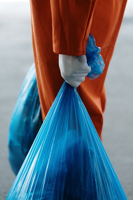 Close-up view of a person wearing orange protective overalls and a white glove, holding a blue garbage bag filled with waste material. The person's hand is wrapped around the tied top of the plastic bag, with the bag's material appearing transparent and slightly crinkled, revealing some of the waste inside. The background is blurred but suggests an outdoor environment, possibly a pavement or driveway, indicating an outdoor rubbish removal activity. The scene highlights the practical aspect of waste collection, with the individual preparing to transport the rubbish, fitting into the context of private waste disposal or on-site clearance provided by companies like Rubbish Removal Bow. The focus on the person's hand and the bag emphasizes manual waste handling, reflecting aspects of independent collection or non-local-authority rubbish management services.