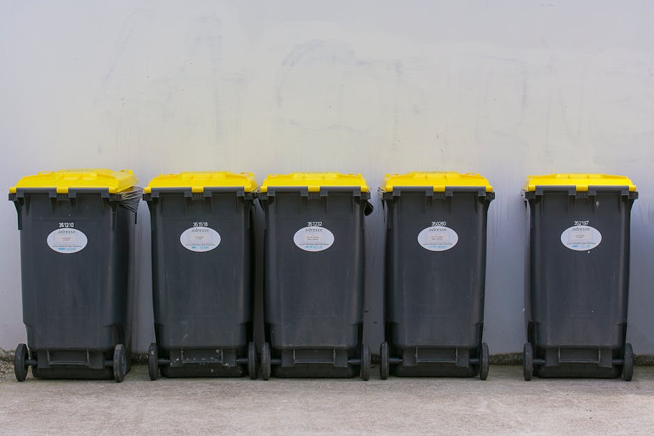 Five large wheeled rubbish bins lined up on a concrete pavement against a plain, light-colored wall. The bins are made of dark grey plastic with textured surfaces and are fitted with bright yellow lids. Each bin features a white oval label with black text and barcode, positioned on the front face. The bins are evenly spaced, with some visible slight scuff marks and dirt on their surfaces, indicating prior use. The scene appears to be in an outdoor urban or residential setting, suitable for private waste collection or alternative rubbish disposal methods. The absence of surrounding elements focuses attention solely on the bins, implying an organized collection point, potentially managed by a waste removal service like Rubbish Removal Bow, specializing in rubbish and waste management services.