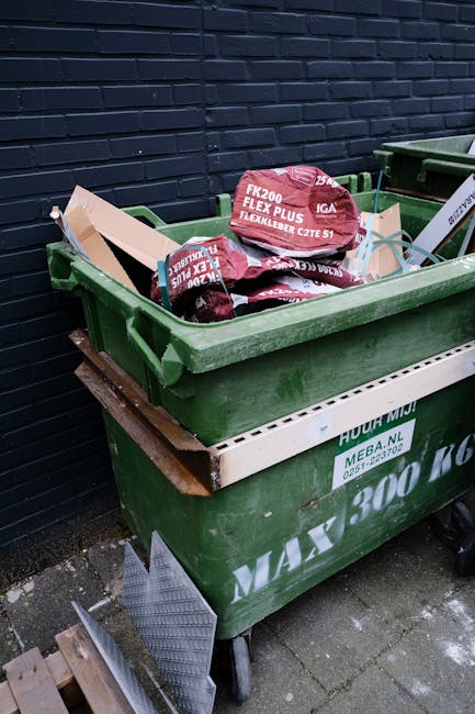 A large green wheeled rubbish bin filled with red and black plastic bags containing building or gardening waste, situated outdoors against a dark brick wall on a paved surface. The bin is partially supported by a white wooden or plastic rail on a wooden platform or pallet, with a partial view of another similar bin to the right. The contents appear to include flexible plastic sheeting or similar materials, indicative of private waste collection or on-site clearance activities. The environment suggests an urban or residential area where waste is temporarily stored prior to collection, aligning with independent rubbish removal services. The scene emphasizes the practical aspect of waste disposal, with visible materials and equipment typical for rubbish removal companies like Rubbish Removal Bow.